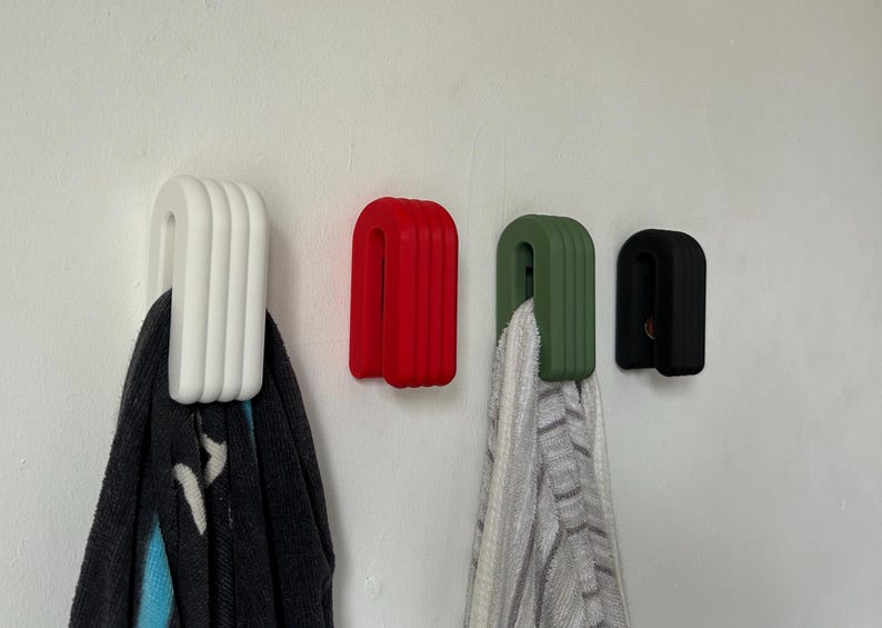 May include: Four wall hooks in white, red, green, and black. Each hook has a rounded, ribbed design. A dark scarf hangs from the white hook, and a striped towel hangs from the green hook. The hooks are mounted on a white wall.