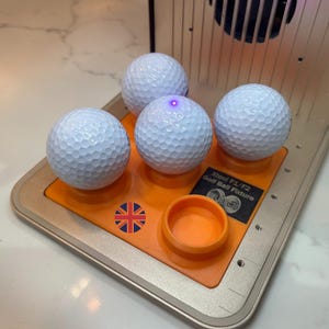 Op de afbeelding: Vier witte golfballen liggen op een oranje platform met een kleine oranje container. Het platform heeft de tekst "Xtool F1/F2 Golf Ball Fixture" en een Britse vlag. De golfballen hebben een putjesoppervlak.