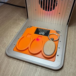 Puede incluir: Una placa de fijación de plástico naranja para máquina láser xTool F1/F2, con tres ranuras ovaladas para llaveros de 50mm x 30mm. Un llavero ovalado de madera clara con una anilla metálica está colocado en la ranura de la derecha.