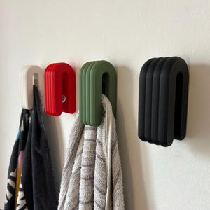 May include: Four U-shaped wall hooks in white, red, green, and black. The hooks are mounted on a white wall, with towels hanging from three of them. The hooks have a ribbed design.