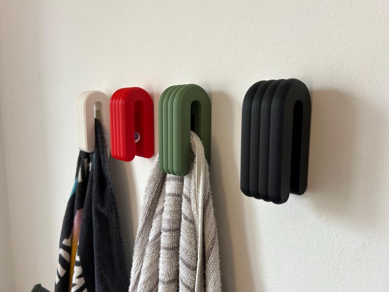 May include: Four U-shaped wall hooks in white, red, green, and black. The hooks are mounted on a white wall, with towels hanging from three of them. The hooks have a ribbed design.