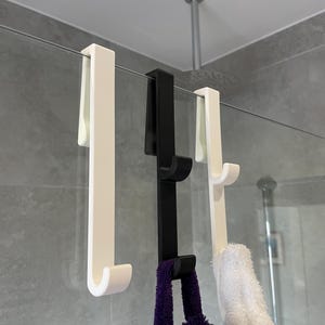 Puede incluir: Tres ganchos de ducha para colgar sobre la puerta, blancos y negros. Los ganchos cuelgan de una puerta de ducha de cristal, con una toalla morada y blanca colgada de los ganchos. Los ganchos son de material resistente.