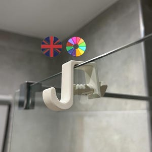 May include: A white, J-shaped shower door hook is attached to a glass panel. The hook is designed to hang items. The background is a light gray wall. Two circular graphics are in the background, one with the Union Jack and the other with a rainbow color wheel.