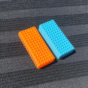 Op de afbeelding: Twee rechthoekige massagetools. Eén is fel oranje, de andere lichtblauw. Beide hebben een textuuroppervlak met kleine, gelijkmatig verdeelde, piramidevormige bobbels. De tools liggen op een donkergrijze, getextureerde ondergrond.