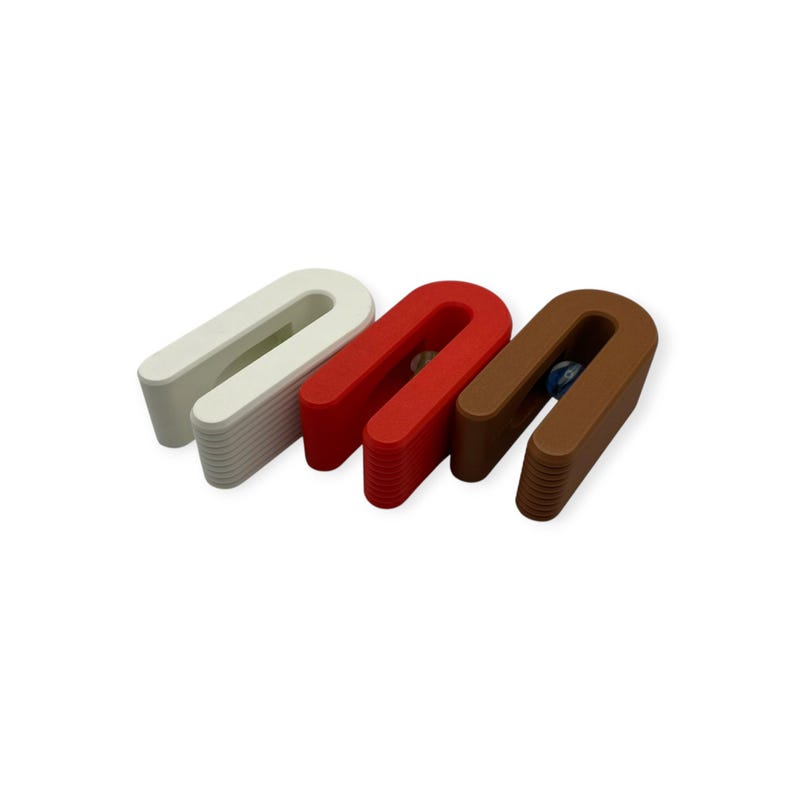 May include: Three U-shaped door stops in white, red, and brown. Each door stop features a smooth, curved top surface and a ribbed, textured base for grip. A small, clear glass marble is visible nestled within the centre of the red and brown units.
