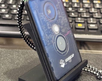 May include: A black and blue electronic device with a circuit board design is displayed on a black stand. The device has a black and white lanyard. The text "SENSECAP" is visible on the device.