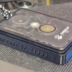 May include: A rectangular black electronic device with the text "Sensecap T1000-E" embossed on the side. The transparent top reveals internal components, including circular elements and the "Sensecap" logo.