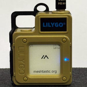 May include: A small, tan-colored device with a black case and a black antenna. The device has a blue screen with the text "meshcastic.org" and a white symbol that looks like a capital "A". The device also has a blue LED light on the right side. The text "Lilygo" is printed on the front of the device.