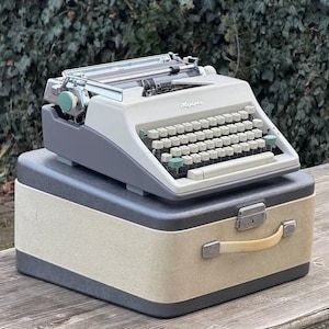 May include: A vintage Olympia typewriter in light grey and dark grey, with a matching carrying case. The typewriter has a full keyboard with white keys and green accents. The case is beige with dark grey trim and a handle.