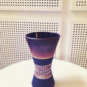 May include: A dark blue vase with a flared top and a decorative band. The vase has a purple and brown band near the top and a patterned band in the middle. The vase is on a white surface.