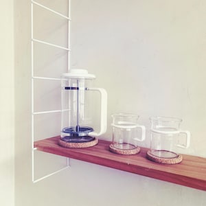 May include: A clear glass cafetière and two glass mugs with white handles are on a wooden shelf. The coffee maker and mugs sit on cork coasters. A white metal shelf unit is mounted on the wall.
