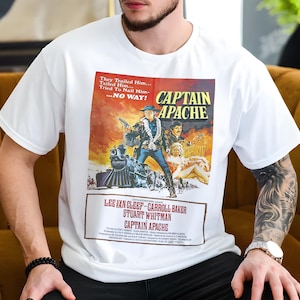 May include: White t-shirt with a vintage "Captain Apache" movie poster design. The poster has a Western theme, featuring a man with a gun, a train, and the film's title. The shirt's design includes the actors' names and the movie title.