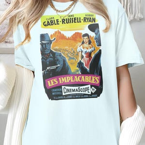 May include: Light blue t-shirt featuring a vintage movie poster for "Les Implacables" with Clark Gable, Jane Russell, and Robert Ryan. The poster has a western theme with a cowboy and woman in a desert landscape. The shirt is a light blue color.
