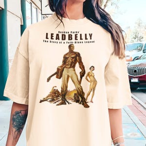 May include: Cream-colored t-shirt featuring a vintage-style illustration of a man with a guitar, a woman, and the text "Gordon Parks' LEADBELLY The Story of a Folk-Blues Legend". The shirt has a relaxed fit.