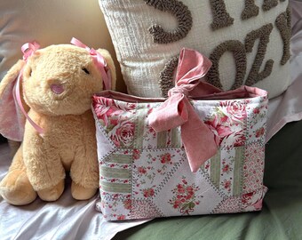 May include: A patchwork fabric bag with a pink bow and floral print. The bag is sitting on a green bedspread with a stuffed bunny and a pillow that says "Stay Cozy".