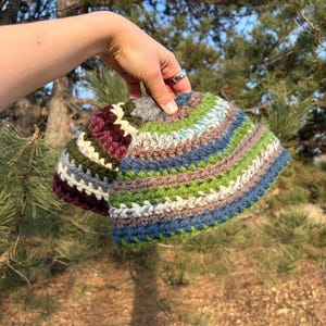 May include: A crochet bucket hat featuring horizontal stripes in shades of forest green, slate blue, tan, cream, and deep burgundy. The hat is constructed with a textured yarn stitch pattern, showcasing a rounded crown and a slightly flared brim.