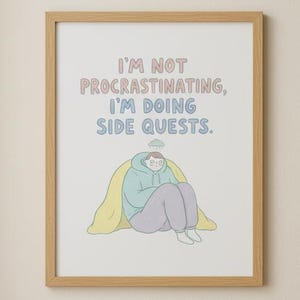 May include: A framed print with the text "I'M NOT PROCRASTINATING, I'M DOING SIDE QUESTS." features a cartoon figure. The print hangs above a green ottoman with pillows and a small wooden side table with a book and mug.