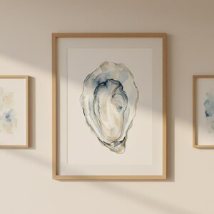 Peut inclure: Trois impressions d'art encadrées à l'aquarelle. La plus grande présente une coquille d'huître détaillée dans des tons de bleu, beige et blanc. Deux petites impressions affichent des motifs abstraits à l'aquarelle bleue et beige. Le tout est dans des cadres en bois clair, créant un thème côtier.