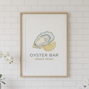 May include: A framed print with an oyster and lemon illustration, labeled "OYSTER BAR" and "SHUCK YEAH!". The artwork is in a light wood frame, hanging above a wooden cabinet with a white countertop. A small orchid and rolled towels are also visible.