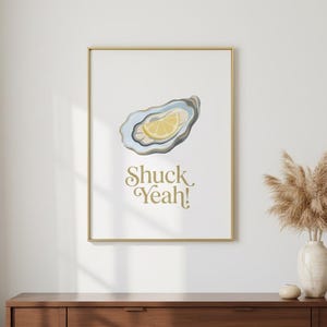 Może przedstawiać: Oprawiony wydruk z ostrygą i plasterkiem cytryny w odcieniach niebieskiego, szarego i żółtego. Wydruk zawiera napis "Shuck Yeah!" złotymi literami. Grafika jest eksponowana na białej ścianie nad drewnianą komodą.