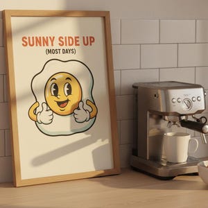 Peut inclure: Une affiche encadrée représentant un œuf au plat de dessin animé avec un visage souriant et les pouces levés, avec le texte SUNNY SIDE UP (MOST DAYS) en lettres rouges et noires sur fond crème, dans un cadre en bois clair.