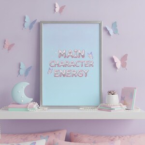 May include: A rectangular silver-framed wall art print featuring the text MAIN CHARACTER ENERGY in a metallic pink balloon-style font on a light blue background, accented with small butterflies, displayed on a white shelf with decor.