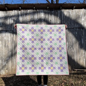 May include: A white quilt with a pink border and a repeating pattern of purple and yellow flowers. The quilt is being held up in front of a wooden wall.