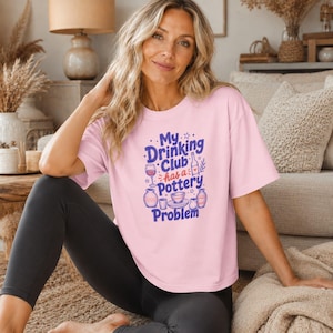 Op de afbeelding: Een lichtroze T-shirt met korte mouwen en een grafische print in paars en rood. De tekst luidt My Drinking Club has a Pottery Problem, vergezeld van illustraties van een wijnglas, een fles, een kom en diverse aardewerken voorwerpen.