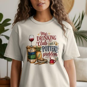 Op de afbeelding: Een crèmekleurig t-shirt met een grafische illustratie van een houten plank gestapeld met kleurrijke keramische kommen, een wijnglas gevuld met rode wijn en een kleine rode mok. De tekst luidt, My DRINKING Club HAS A POTTERY problem.