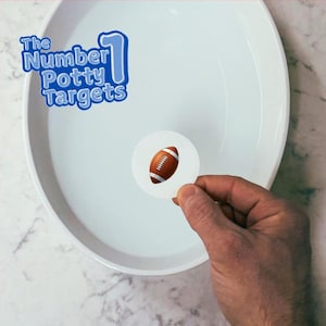 May include: A white bowl with water and a round sticker with a brown and white football on it. The sticker is being held by a hand. The text "The Number 1 Potty Targets" is visible in the top left corner of the image.