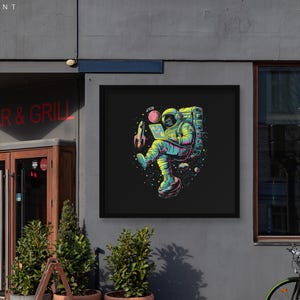 May include: A framed art print showing a vibrant illustration of an astronaut working on a laptop in space. The astronaut, in a yellow and blue spacesuit, is surrounded by planets, a rocket, and stars. The artwork is displayed on a grey building exterior.