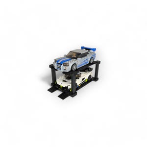 Puede incluir: Un elevador de coches LEGO negro con dos coches encima. El coche de arriba es un coche deportivo plateado y azul. El coche de abajo es un coche deportivo blanco.