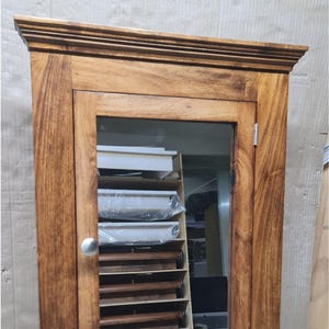 Könnte beinhalten: Ein hölzerner Medizinschrank mit Spiegeltür und silbernem Knopf. Der Schrank hat eine warme braune Oberfläche und verfügt über eine Reihe von Regalen, die durch das Glas sichtbar sind. Das Design ist traditionell und funktional.