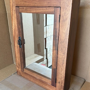 May include: A rectangular wooden medicine cabinet with a mirrored door. The cabinet is stained a warm brown color, with visible wood grain. It features a black metal handle and hinges. The cabinet is designed to be mounted on a wall.