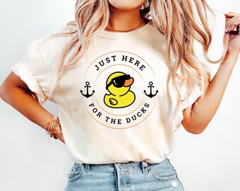 May include: Cream-colored t-shirt with a circular graphic featuring a yellow duck wearing sunglasses. The text "JUST HERE FOR THE DUCKS" surrounds the duck, with anchors on either side. The shirt has short sleeves.