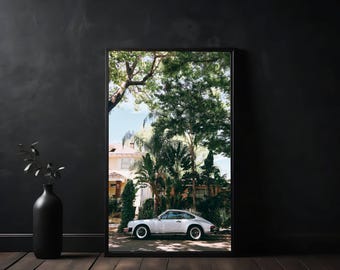May include: A white vintage car parked in front of a house with a lush green lawn and trees. The car is facing the camera and has a white body with black trim. The car is parked on a paved driveway.