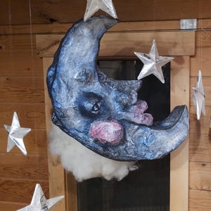 May include: A whimsical, handcrafted moon and stars mobile. The moon is a crescent shape with a blue and gray textured surface, pink cheeks, and a cloud of white fluff. Silver stars hang from strings.