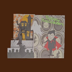 May include: A Halloween-themed papercraft card featuring a haunted house, a vampire, a ghost, and the text "Happy Halloween." The card has a layered design with orange, gray, and white elements, and a brown background.