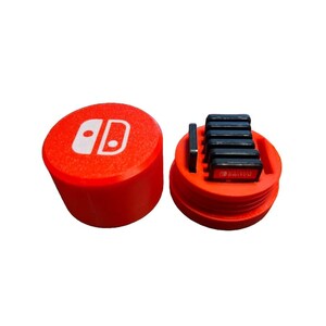 May include: A red, cylindrical case with a white Nintendo Switch logo on the lid. The case is open, showing several black game cartridges inside. The case is designed to store and protect game cartridges.