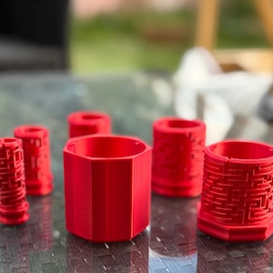May include: Several red 3D-printed containers are displayed. The collection includes cylindrical and hexagonal shapes, some with detailed patterns. The objects are arranged on a reflective surface, showcasing their design.