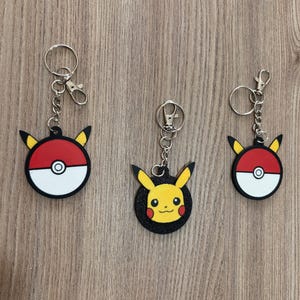 May include: Three keychains featuring Pokemon characters. The first keychain is a black and red Pokeball with a yellow star. The second keychain is a black and yellow Pikachu with a red cheek. The third keychain is a black and red Pokeball with a yellow star.