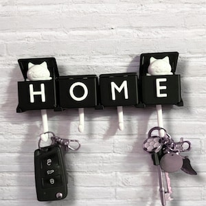 May include: A black key holder with the word "HOME" in white letters. Each letter has a white cat figurine on top. Keys and a car remote hang from the hooks below. The key holder is mounted on a white brick wall.