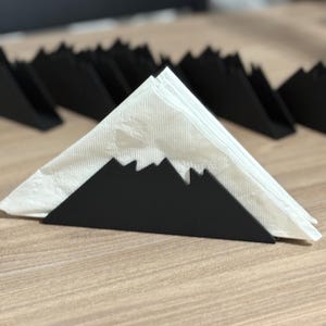 May include: Black metal napkin holder shaped like a mountain range with a white paper napkin inside.