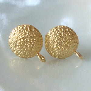 Puede incluir: Un par de pendientes de color dorado con un acabado texturizado martillado. Los pendientes son redondos y tienen un diseño simple y minimalista.