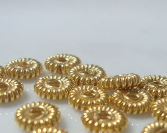 May include: A group of gold-colored metal beads with a decorative, twisted design. The beads are arranged in a random pattern on a white surface.
