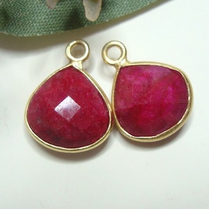 May include: Two gold-plated pendant charms with a teardrop-shaped ruby gemstone. The ruby is a deep red color and has a faceted cut.