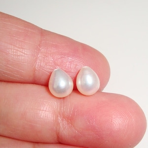 May include: Two white pearlescent teardrop-shaped beads, approximately 8mm in size, are shown on a finger.