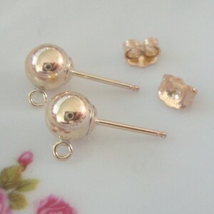 May include: Two gold-toned ball earring posts with a loop on the top of each ball. The earring posts are on a white surface.