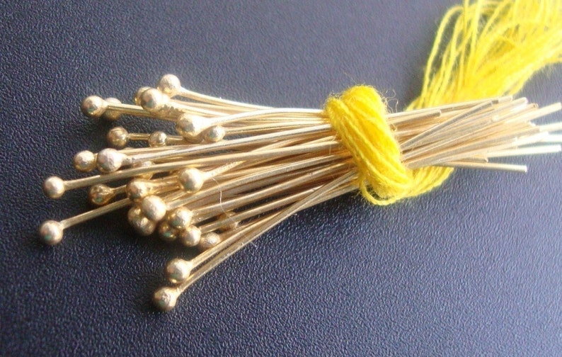 Puede incluir: Un conjunto de alfileres dorados con cabezas redondas, atados con hilo amarillo. Los alfileres se presentan sobre una superficie texturizada azul oscuro, resaltando su brillo met&aacute;lico y su dise&ntilde;o delicado.