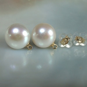 May include: Two large, round, white pearl earrings with gold-colored loops, next to two clear, flower-shaped earring backs. The pearls have a subtle iridescent sheen. The earrings are set against a light, neutral background.